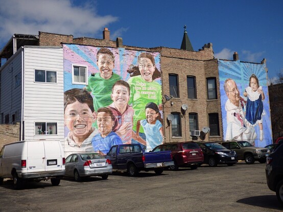 rear elevation of an apartment block, filled with the happy faces of active children, and a priest holding up in a blue velvet dress with lace collar.  a young girl