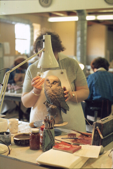 A pottery owl is standing on a turn table, in it's raw clay form; it's being worked on by a craftsperson, whose face is blocked by a classic anglepoise lamp.  Around it are paint brushes, materials, and an old mucky radio.  The owl is superb.