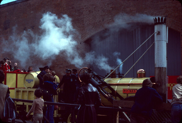 A yellow replica of the Rocket steam train puffs out steam.