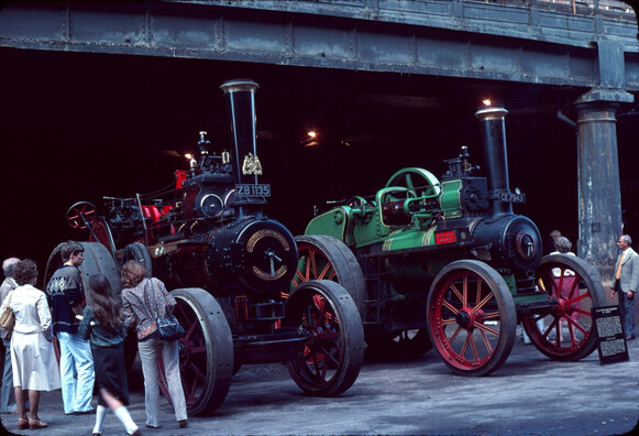Two large general purpose steam engines sit on the road as people look at them.