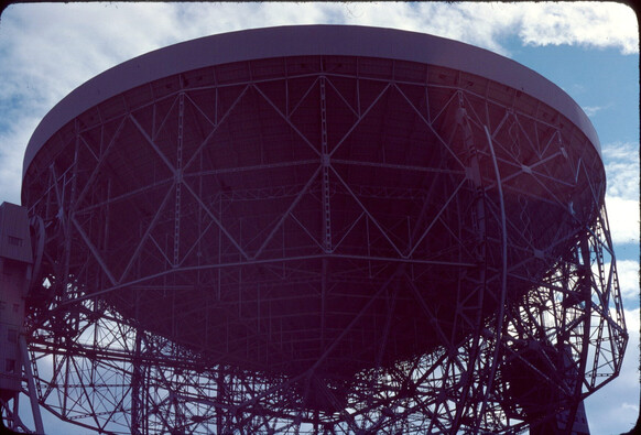 A large steel-frame built radio telescope facing skywards.  A large network of steel trelliswork holds it's bowl up.  There are some fluffy clouds above.