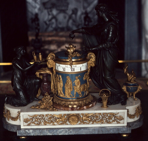 An ornate cylindrical clock. The clock itself is a cylinger with bands at the top (I think they rotate).  It's decorated heavily in gold.   I'ts on a white and then grey marble base, more gold.  It's surrounded by two black brass figures.  Very very ornate.
