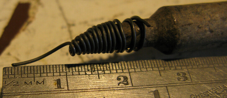 A close up of a soldering iron bit with a piece of wire tightly wrapped around it with a small tail;  it's next to a metal ruler showing the size of the wire