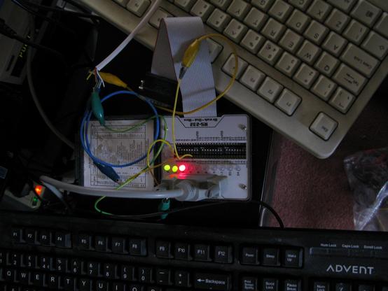 A horribly hacky serial setup; between two keyboards sits a serial breakout box, various wires and crock clip leads.  It's working by good luck rather than physics, The lights for RX/TX are green and the next LED is red.
