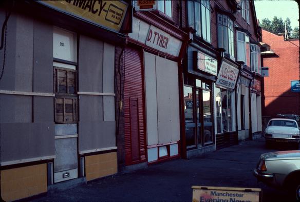 A small row of shops in the early 1980s, two of the shops have makeshift wooden barriers over the fronts.   It's a fairly basic set of small UK shops, with windows over the top.