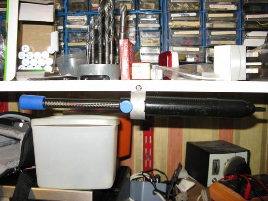 A huge solder sucker hangs from a misused valve clip screwed to a shelf.  The shelves above and below have a miscellany or random workshop stuff; components in drawers, drills, batteries, plugin testers, small electronic things.  Damn it's a big solder sucker.