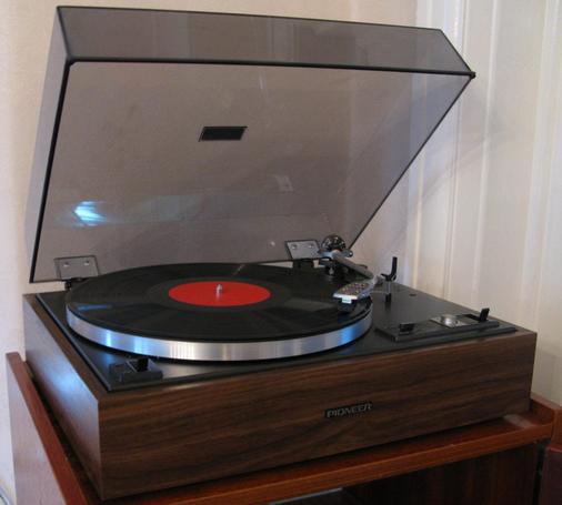 A record turntable, a pioneer PL-15R, with a record playing.  The player has 70s faux wood as it's surround, and the player itself has a black base, and black and silver controls.  On top of a silver platter is a record with a bright red label.  A translucent lid is hinged upwards. 