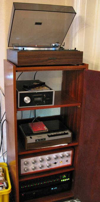 A hifi cabinet, it's about 5ft tall, and probably 3ft ish wide.  It's just wide enough for the record player (in the other pic) to sit on top.  The cabinet is a dark faux wood, with an open door.  Inside there's a radio, a cassette deck, an amplifier and a graphics equalizer. A light glows on the amplifier, and the graphics equalizer has many lights on it's sliders and a frequency display.