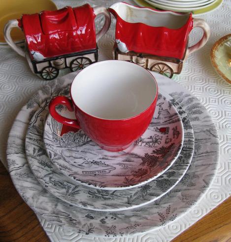 A tea cup, bright red on the outside, white inside, sits on saucer, side plate and full plate which are mostly black and white patterned; in the background is a milk jug and hmm, sugar bowl? whicha re done as covered wagons in red and cream.