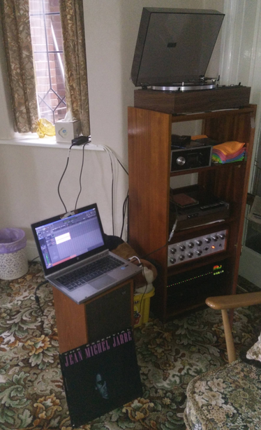 A laptop is sat ontop of a small speaker next to a rack of mostly old hifi stuff including a turntable on top.  In front is the cover of Jean Michel Jarre's  'Essential'.  The laptop is displaying Ardour's screen.