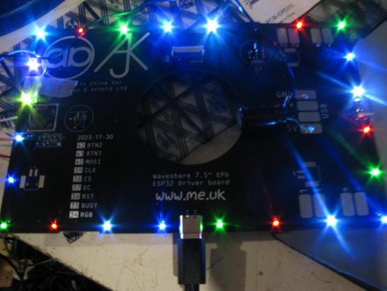 a PCB with a large circular hole in it sits on a bench with a USB-C cable coming out the bottom and various wires; around the edge are a set of bright LEDs in different colours.  They all have lens flare.  Did I say they're BRIGHT.