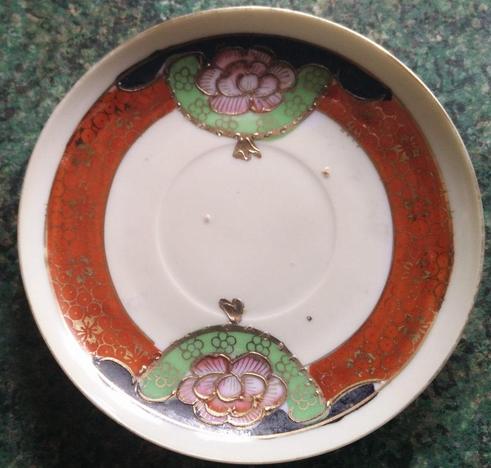 A small saucer; it's mostly white with a red/orange rind around and a couple of pink flowers surrounded by some green; there's some golden decoration on top.