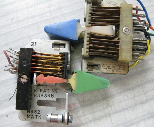 A pair of old toggle switches sitting on a table.  They've got a metal frame and then sets of (exposed) leaf switch contacts that are pressed or released by the tilt of the central column that moves based on the toggle at the front.  They have bits of wire soldered on where they were once used.  
One is marked 'Made By Telephone Mfg Co. Ltd under license from Ericsson Teles Ltd.  Pat No 838348'.The other is marked K. Pat No 838348, N9321 MATK'
One has a pleasing blue toggle, the other a pleasin…