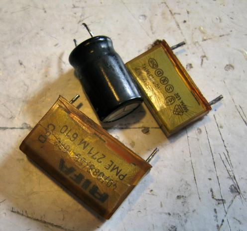 3 capacitors sitting on a white bench.  One is a black cylindrical; the other two are nasty rectangular RIFA caps, discoloured brown with some cracks in the case.