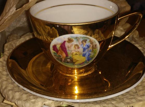 A very blingy tea cup and saucer; the saucer is just shiny gold effect (with white underside); the cup inner is white with a thin gold line, the outer is again shiny gold, with an inset classical panel with 3 ladies in Red, yellow and blue