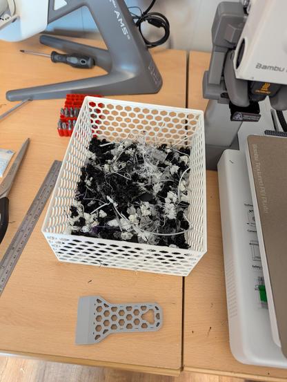 A plastic basket with hexagon shaped holes in the size filled with stringy plastic waste from a 3d printer. It is sat on a bench with a 3d printer just in view on the side.