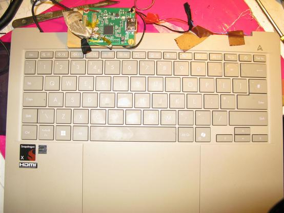 The base of an Asus zenbook Snapdragon laptop, notably missing it's lid/display entirely.  On the top edge are some wires glued on and a small eDP->HDMI converter board mounted on an off cut of wood.  It's sitting on a work bench with some purple folder behind it and a scalpel.