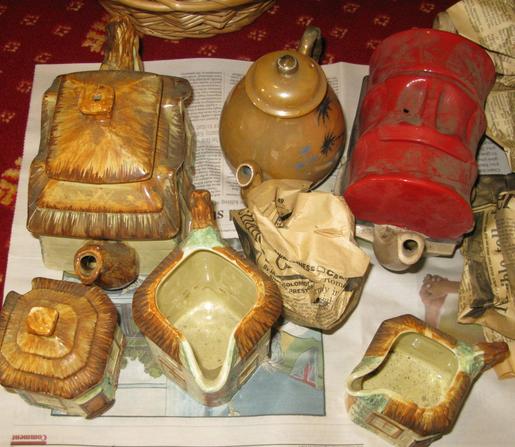 Three very decorative teapots, and some sugar bowls and jugs sit on a piece of paper.  They're covered in dust.  One teapot is made like a rustic farm house with the lid being thatched roof, the next has palm trees on, the third had a red top and is made to look like an old west horse wagon.