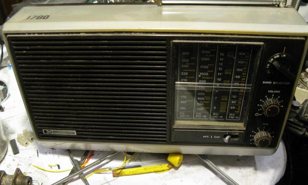 A Pye 1780 portable radio tuned to Radio 4 Long wave, on a  rather messy bench. The radio is from the late 70s, and has a grey case with black speaker grill and frontage, a vertical scale with clear plastic cover over the scale and some knobs with orange highlighting.