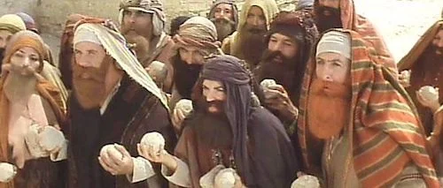 A group of people wearing layered robes and various head coverings are gathered together. The individuals wear fabrics in shades of tan, brown, white, and grey, including turbans and draped cloths. Many of the people have visible facial hair such as beards and mustaches. Several people in the foreground hold small, round, light-colored objects that resemble bread. The background is a bright, neutral color.
