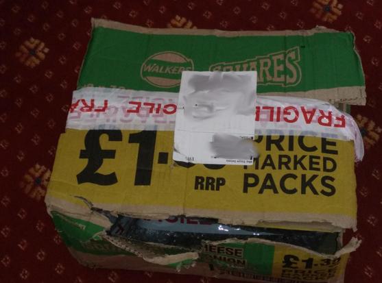 A battered reused crisp box sits on a  red carpeted floor.  It's split along at least one edge.  The main seam is held shut by 'fragile' tape.  The box was for Cheese and Onion Walkers squares.