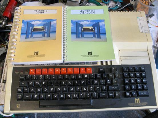 A BBC Master 512 computer - beige case, BBC keyboard with the Red function keys, (but with the arrow keys in a different orientation to a Model B) and a numeric keypad.  Two cartridge slots on the right are empty.   It has two small added switches on the back right.  On top are the 'Welcome Guide' (for a base Master 128) and 'Master 512 user guide'