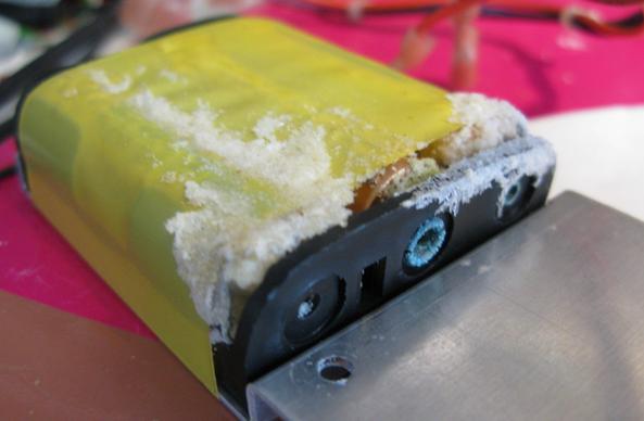 A 3 cell battery pack sits on a bench attached to a bracket with some yellow tape.  There's white furry deposits around it, one of the contacts is very green. They were Duracell batteries.