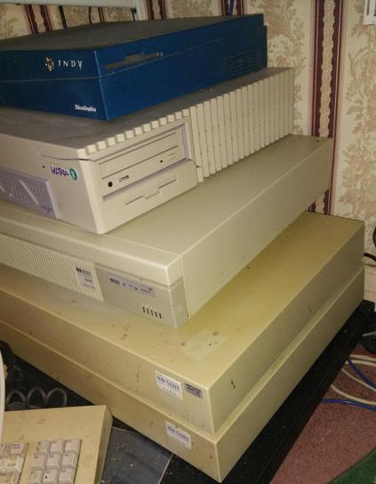A stack of old Unix workstations on a trolly.  From top to bottom; an SGI Indy, a Sun Ultra 1, an HP 9000/735 and two Sun 3/60's.  In front is the corner of a Sun3 optical mouse mat and keyboard.  They're sitting on a black dolly.